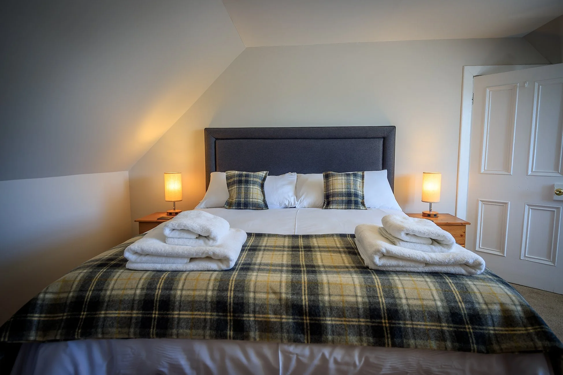 A neatly made bed with plaid bedding, four rolled white towels on top, two plaid pillows, in a cozy bedroom with two bedside lamps, a gray headboard, cream walls, and a white door.