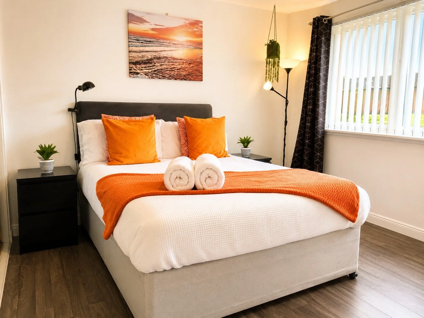 A neatly made bed with white bedding, two orange pillows, and a matching orange blanket at the foot. Two rolled white towels are placed on the bed. There are two black nightstands with small green plants on each side of the bed. A black wall-mounted 