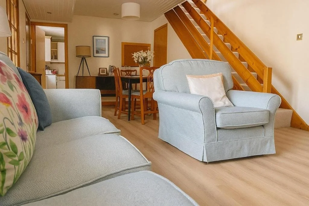A living room with a light blue armchair with a white pillow, a sofa with floral and dark pillows, a wooden staircase, a round wooden dining table with four chairs, and a doorway leading to the kitchen.