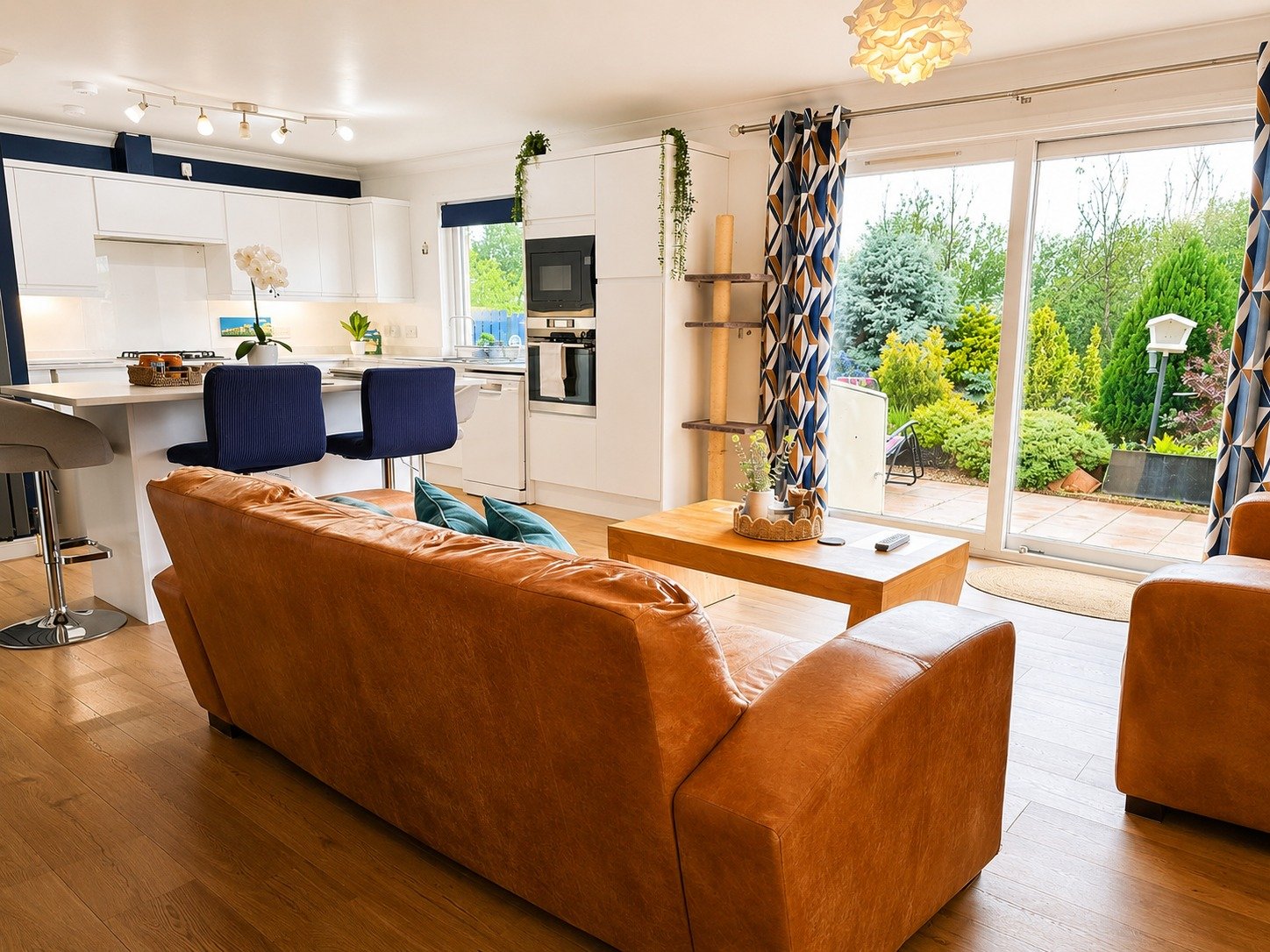 Living room with brown leather sofa, wooden coffee table, and large sliding glass door leading to a garden with lush greenery and outdoor seating