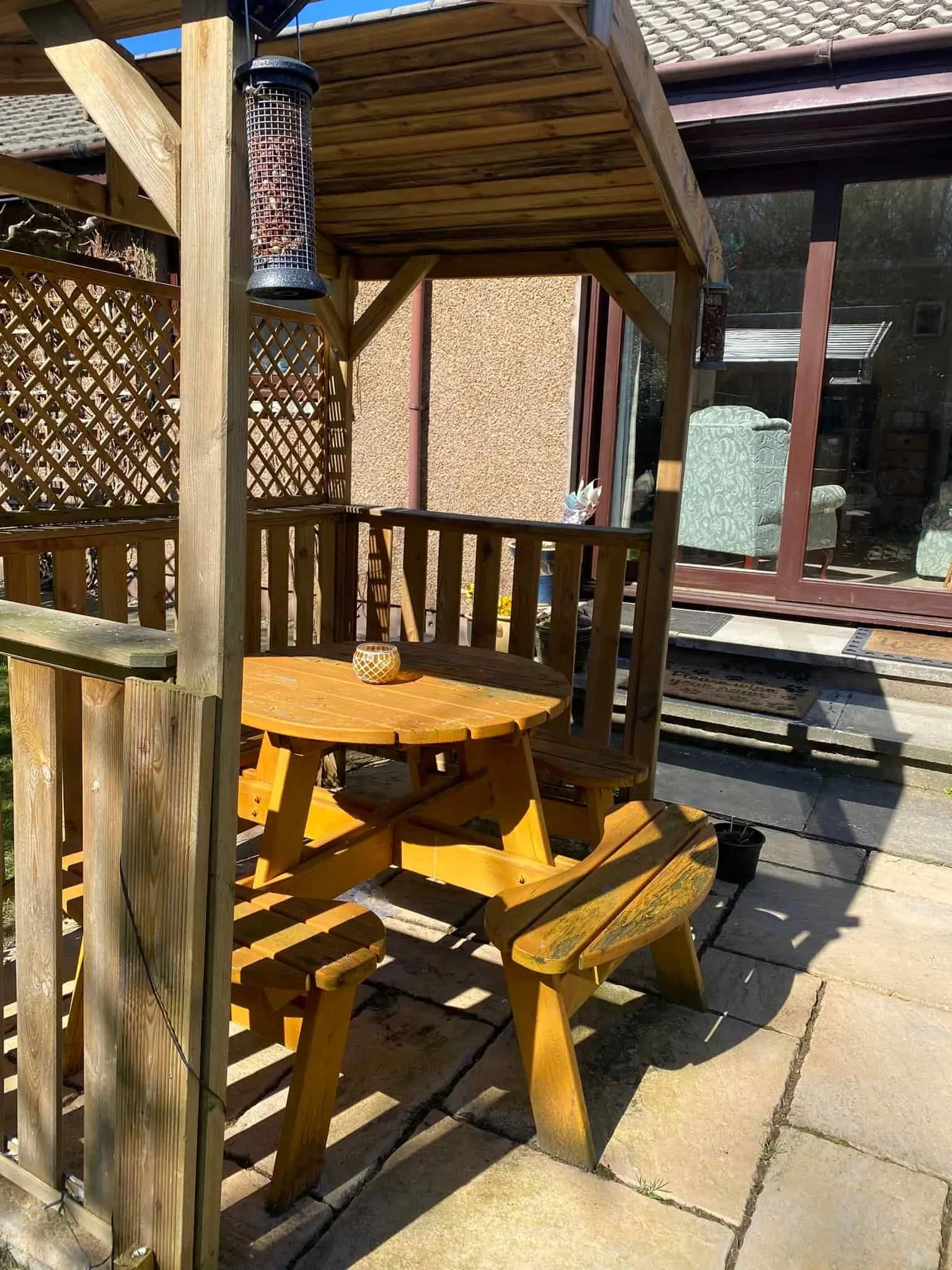A small outdoor wooden gazebo with a round wooden table and attached benches, situated on a stone patio, with a black bird feeder hanging inside and a sliding glass door leading into a house in the background.