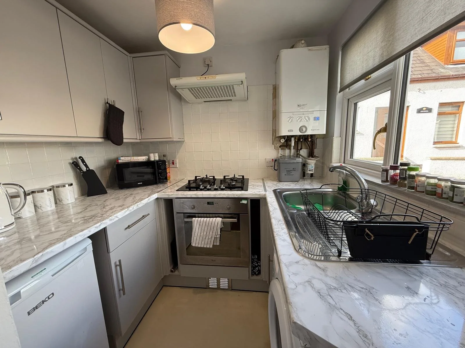 Small kitchen with white cabinets, marble countertops, a stove, microwave, and a sink near a window with kitchen jars on the sill.
