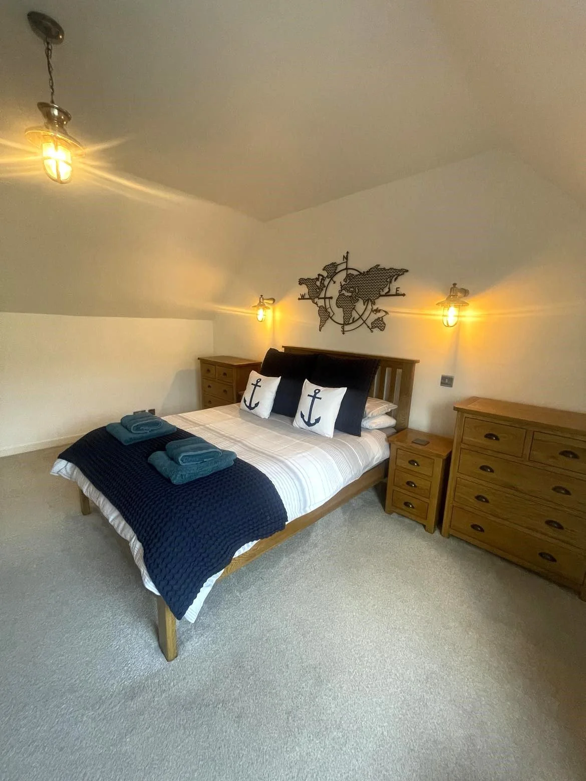 A bedroom with a wooden bed frame, nautical decor, two pillows with anchor designs, and two folded towels, illuminated by wall-mounted lights.