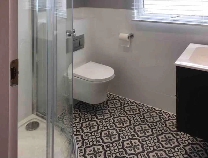 A small bathroom with a corner shower, a wall-mounted toilet, a window with white blinds, and a black vanity with a white sink. The floor has black and white patterned tiles.