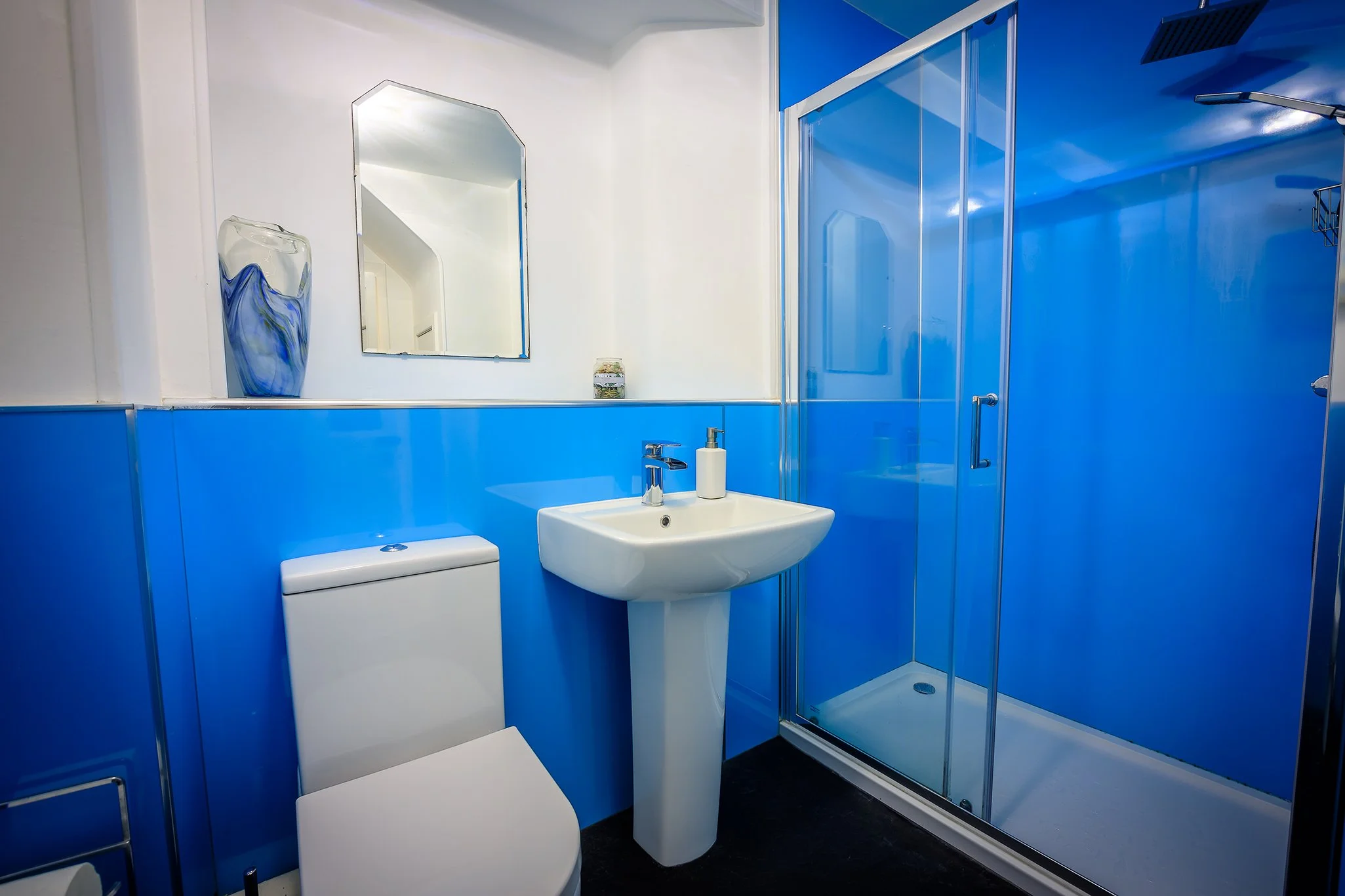 Bathroom with blue walls, a white toilet, a white pedestal sink with a soap dispenser, a mirror, and a glass shower enclosure.