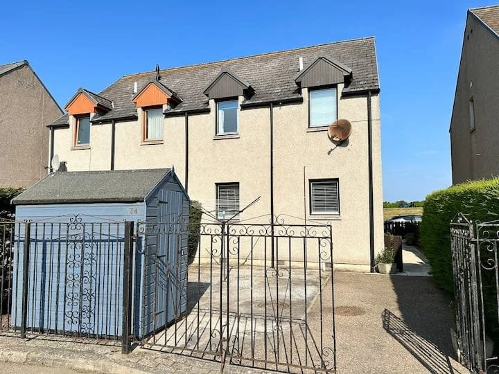 A beige multi-story house with a gray roof, three small roof dormers, and a large satellite dish attached to the wall. A metal gate and fence, along with a blue shed, are in the front yard. There is a pathway leading to a side area with greenery.