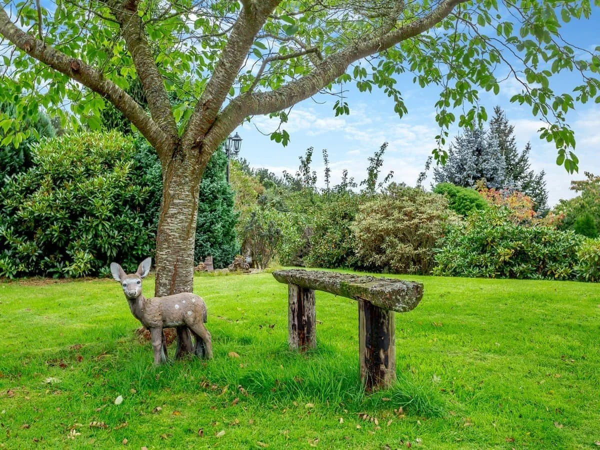 A small deer statue standing on a grassy lawn under a large tree in a garden with bushes and trees in the background.