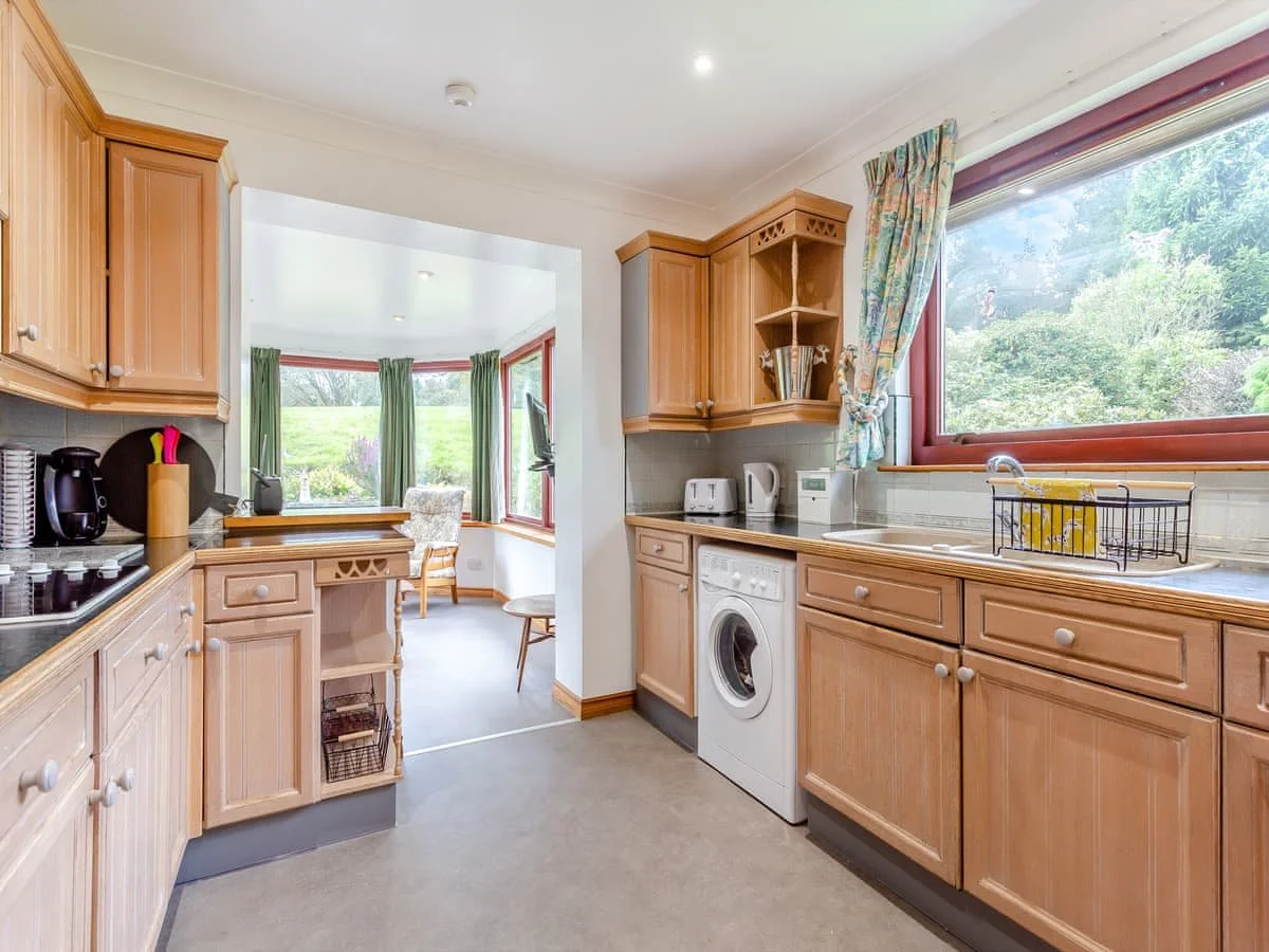 Kitchen with wooden cabinets, a window showing green trees outside, a washing machine, small appliances, and a view into a bright sunroom with large windows and chairs.