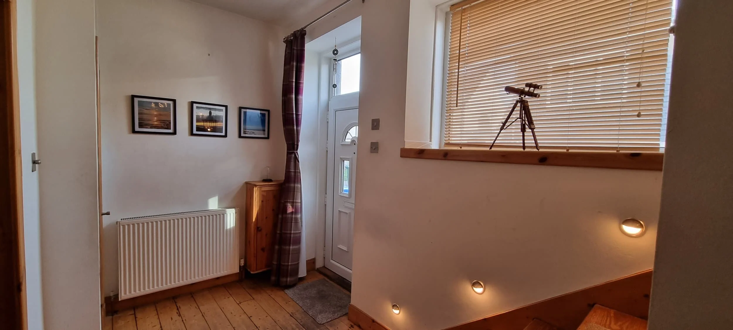 Entryway with a door, a small wooden cabinet, and a wall-mounted radiator. The wall features three framed photos of nature scenes. To the right, a window with closed horizontal blinds and a small telescope on the windowsill. Recessed lighting illumin