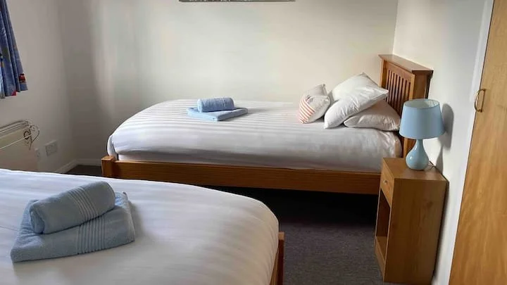 Two single beds with white and striped bedding in a simple bedroom. One bed has a rolled towel on top, the other has pillows and a blanket. There is a wooden nightstand with a blue lamp next to one bed, and a small window with a curtain on the wall.