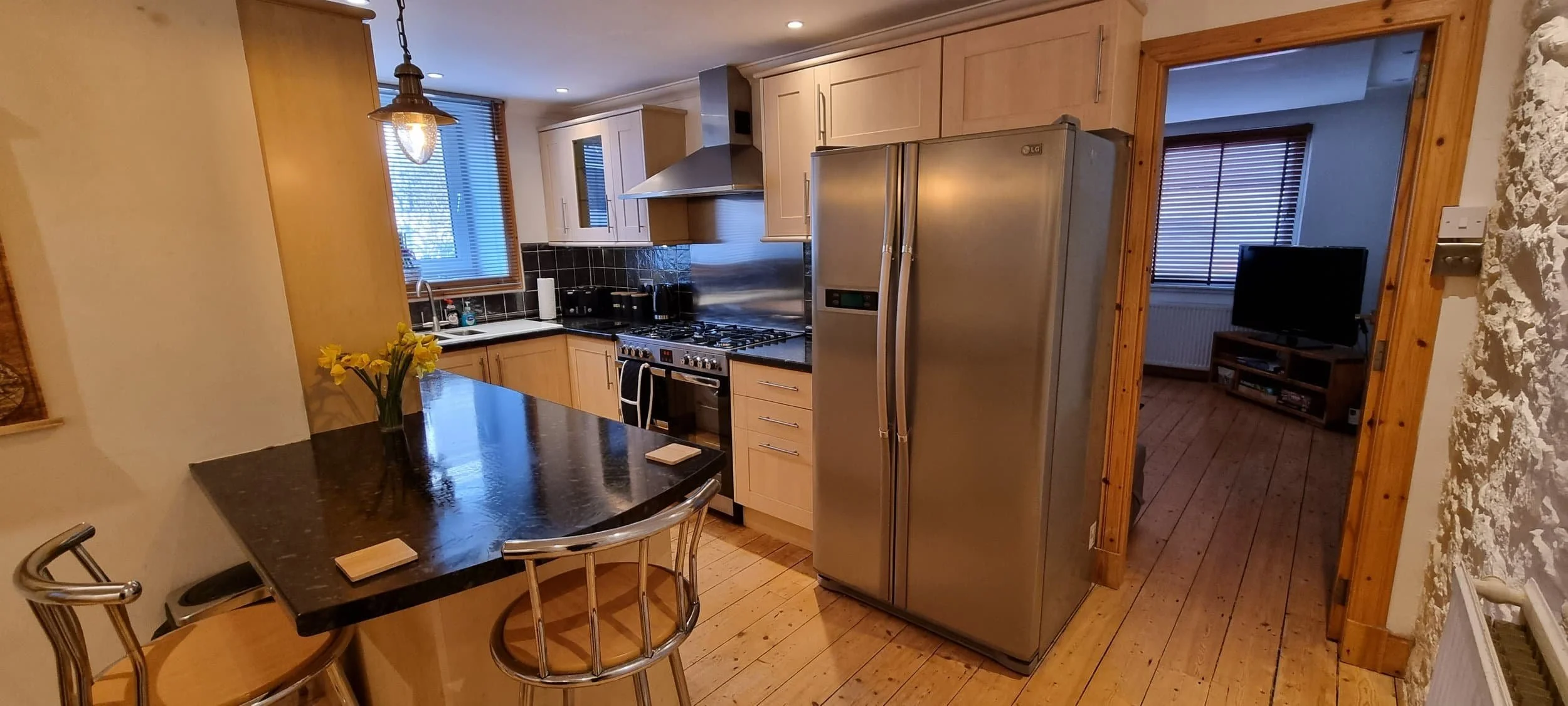 Kitchen with granite countertop island, wooden cabinets, stainless steel appliances, and dining chairs. Adjacent to a living room with a TV, wooden floor, and window with blinds.