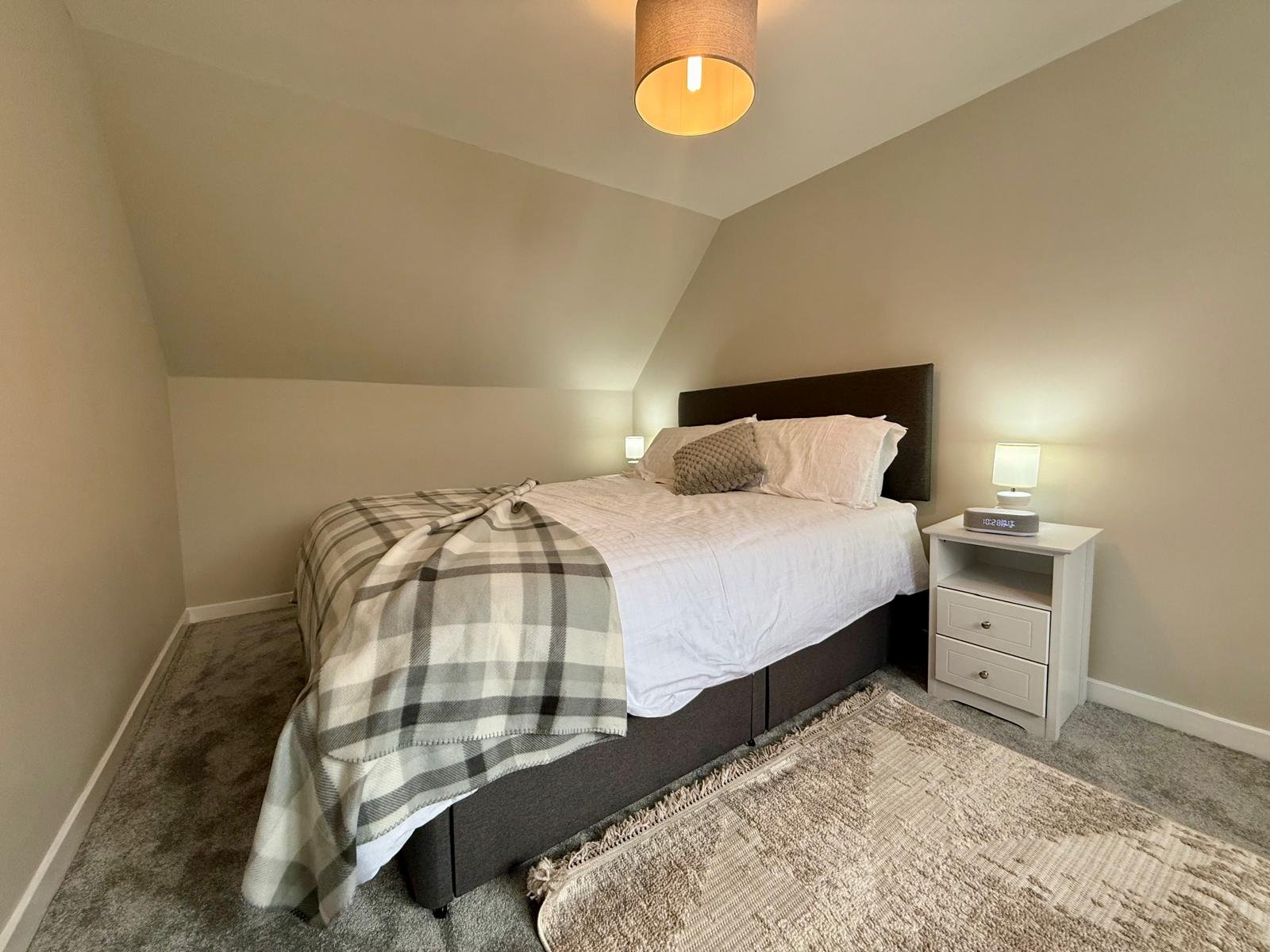 A neatly made bedroom with a queen-sized bed featuring a dark wood headboard, white bedding, and a plaid blanket. There are pillows on the bed, and a small white nightstand with a lamp and an alarm clock. The room has beige walls, a textured rug, and