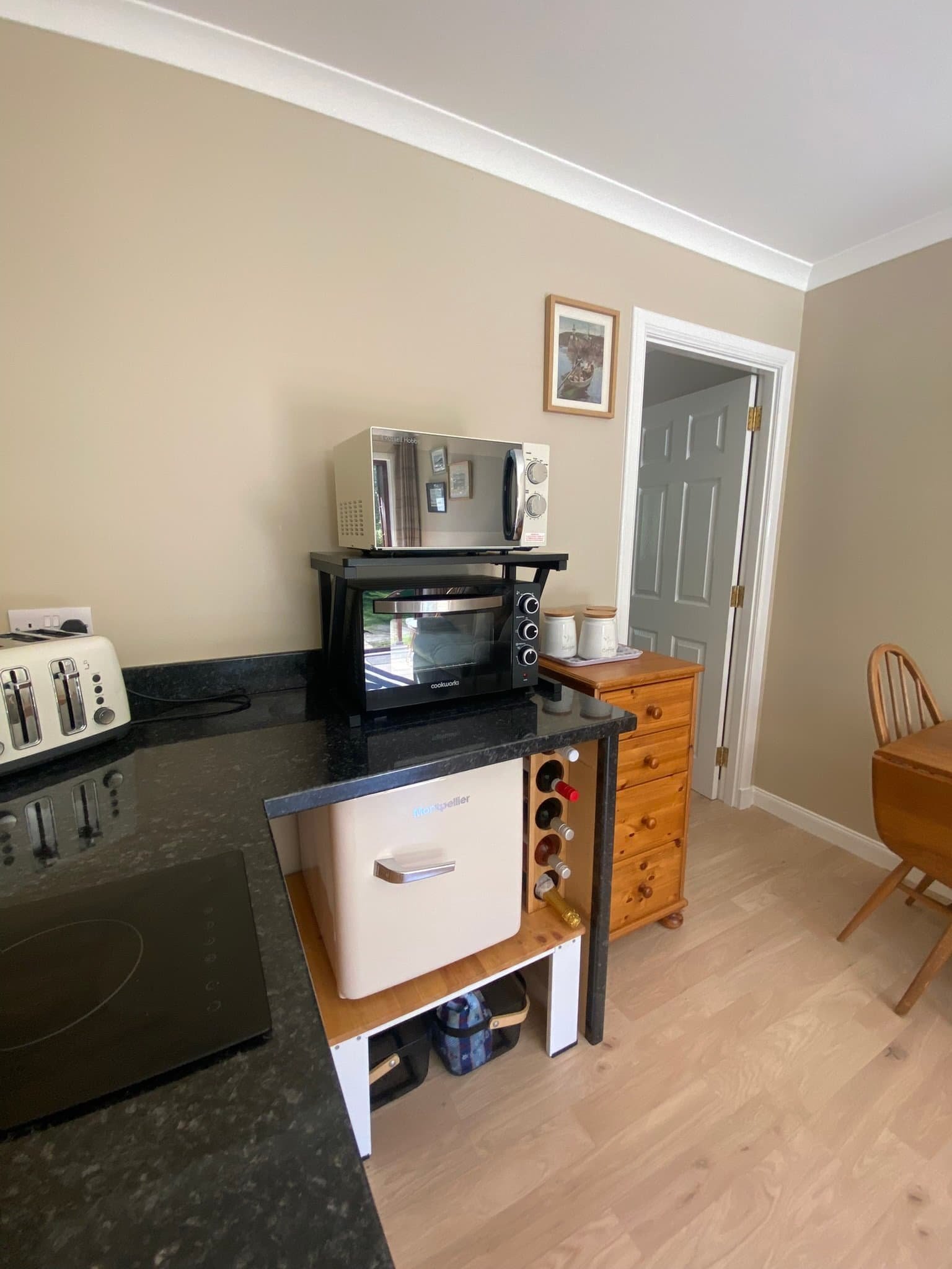 A kitchen corner with a black countertop, small appliances including a toaster and microwave, a wine fridge, a small wooden dresser with three ceramic jars, and a round wooden dining table with chairs. The wall has a framed picture, and there is a do