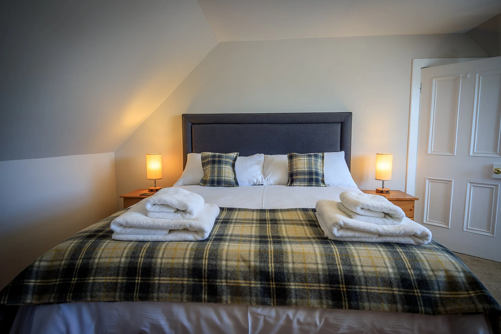 A neatly made bed with a blue headboard, two plaid pillows, and folded towels on top, in a bedroom with two bedside lamps and a white door.