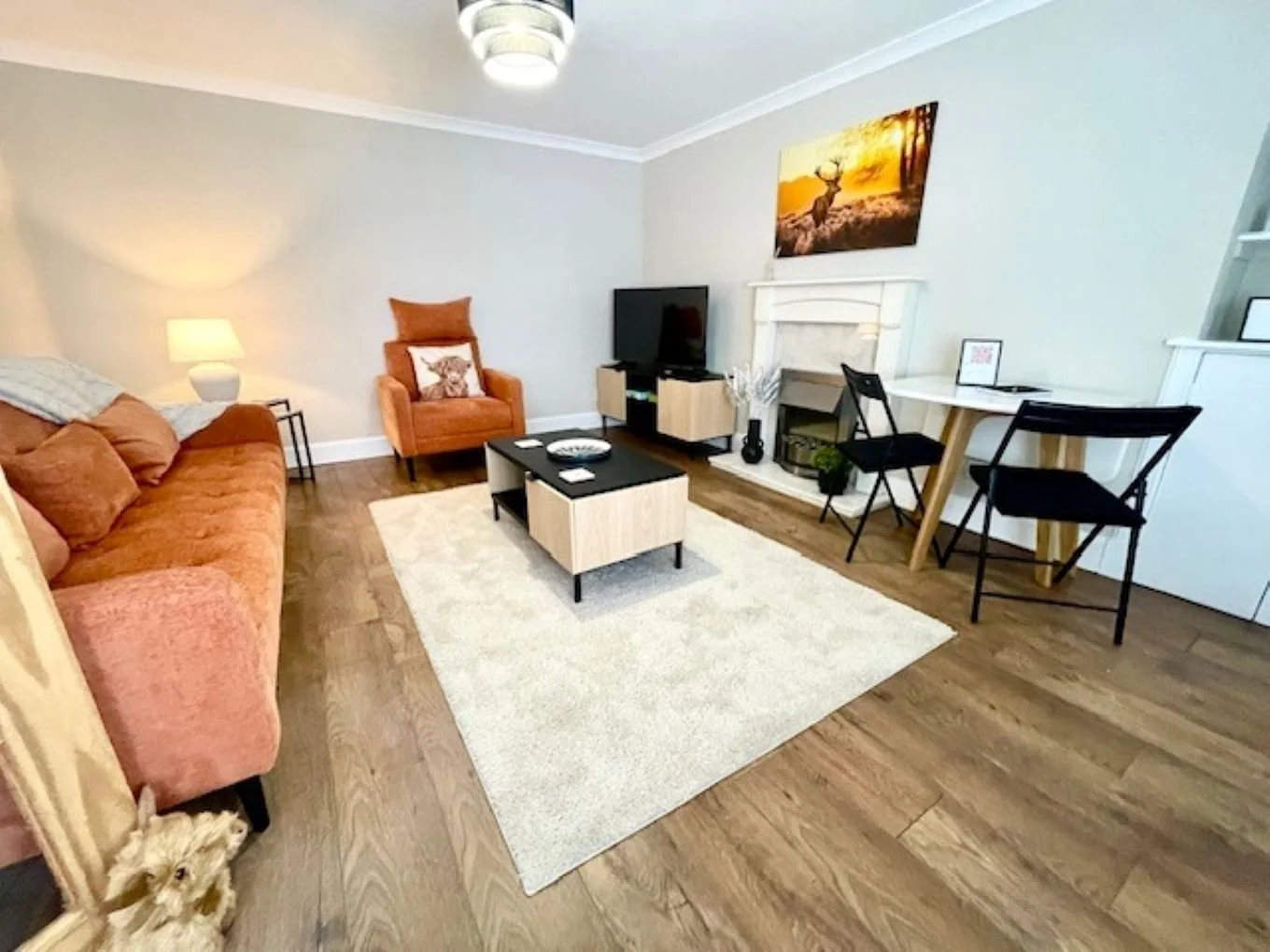 Living room with a coral sofa, an orange armchair with a deer pillow, a white area rug, a black coffee table, a small white TV stand with a TV, a white fireplace with a picture of a deer near a sunset, a white desk with two black chairs, a painting o