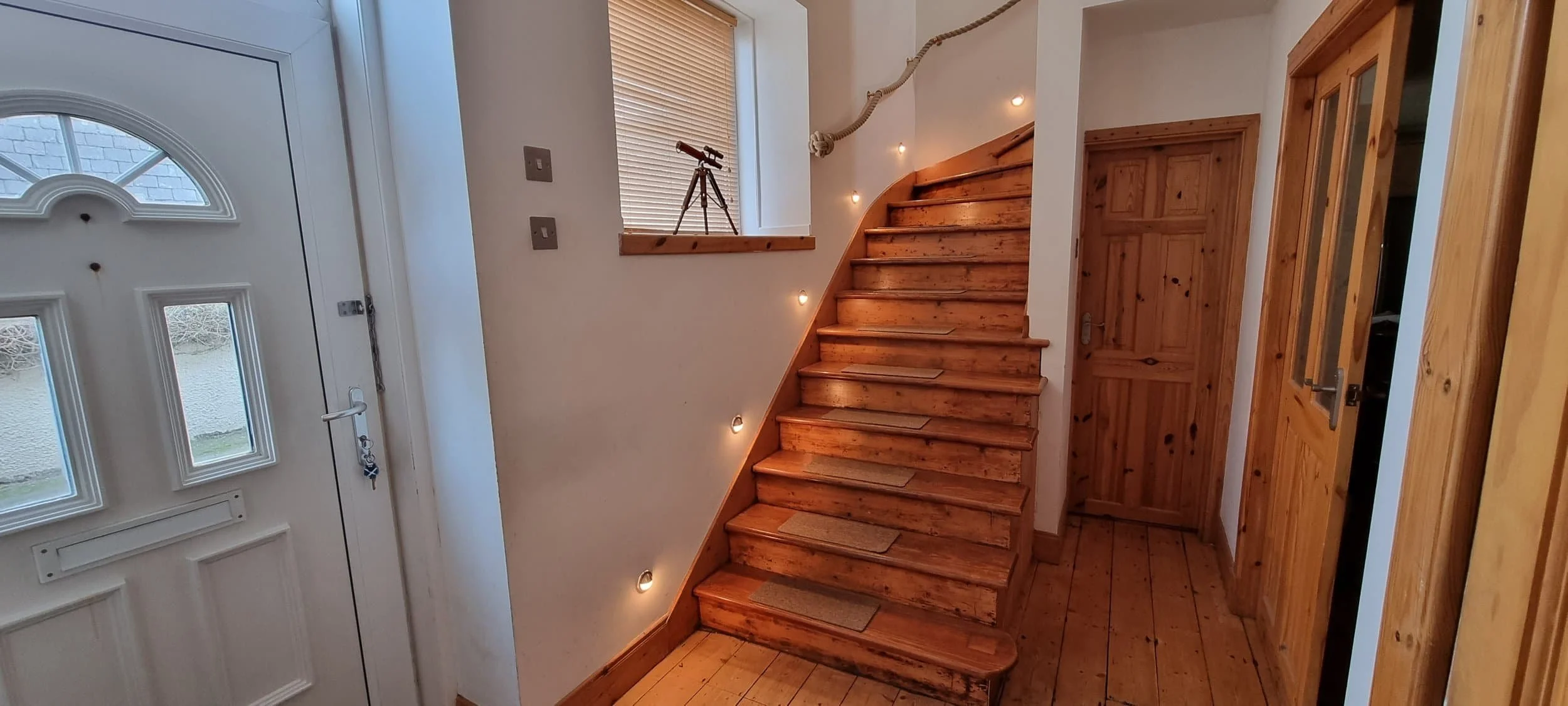 Interior of a house entryway with a white front door, wooden stairs with a rope handrail, a window with blinds, a telescope on the window sill, and wooden doors on the adjacent walls.