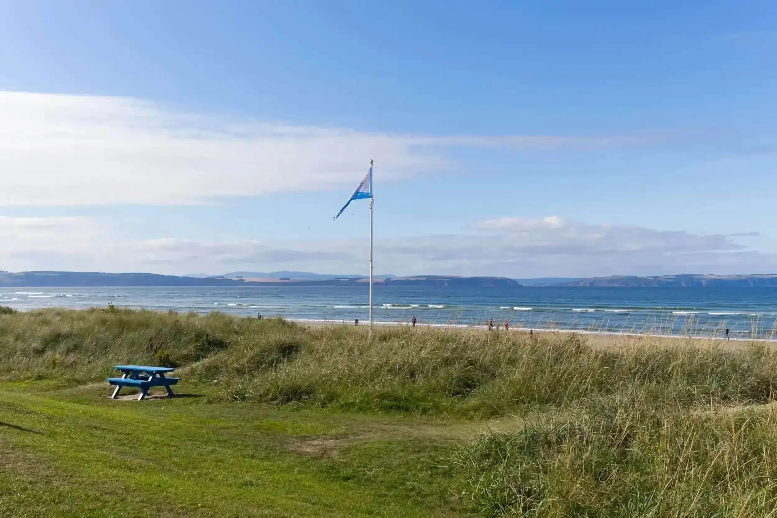 A coastal view with a grassy area, a blue picnic table, a flagpole with a blue and white flag, sandy beach, ocean waves, and distant land across the water under a partly cloudy sky.