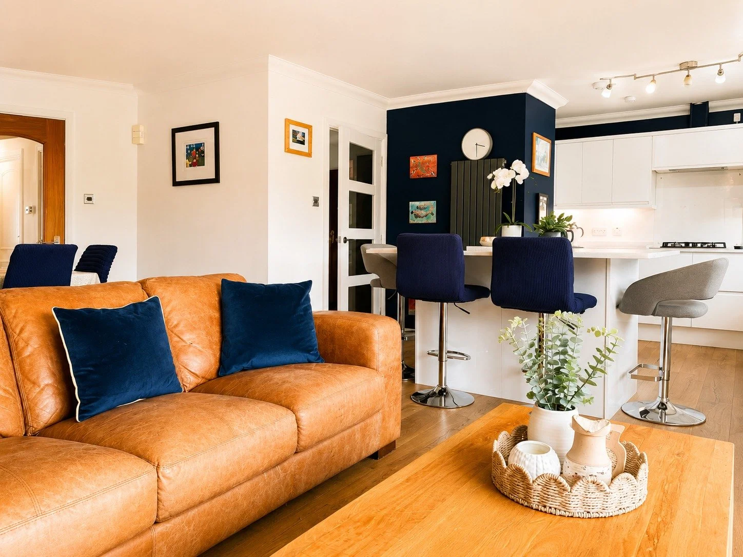 A living room with a tan leather sofa, blue pillows, and a wooden coffee table with a white planter. The background shows a kitchen with a dark blue accent wall, white cabinets, and a kitchen island with four chairs, two blue and two gray. There are 