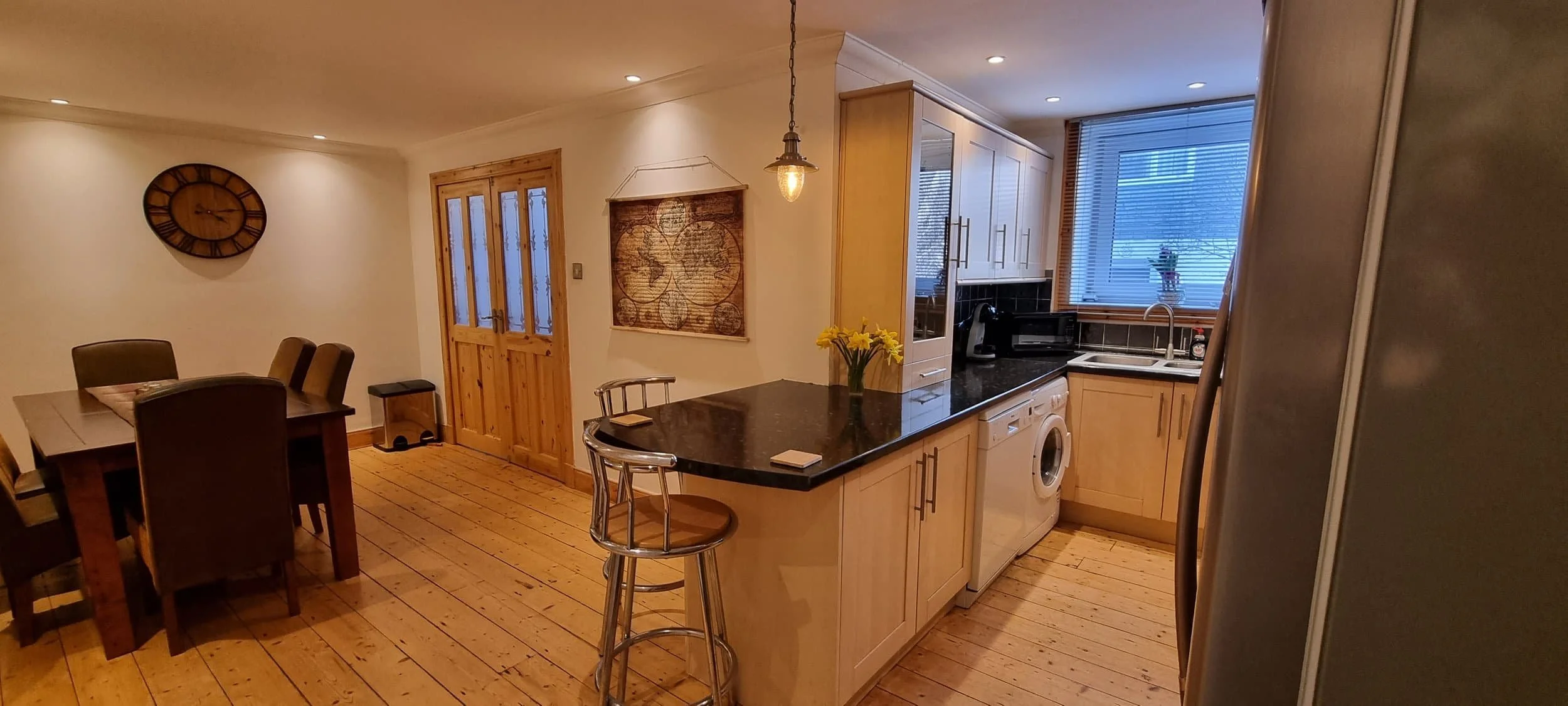 Kitchen and dining area with wooden floors, cream cabinets, black countertops, large window with blinds, dining table with chairs, decorated with flowers, and a wall clock.