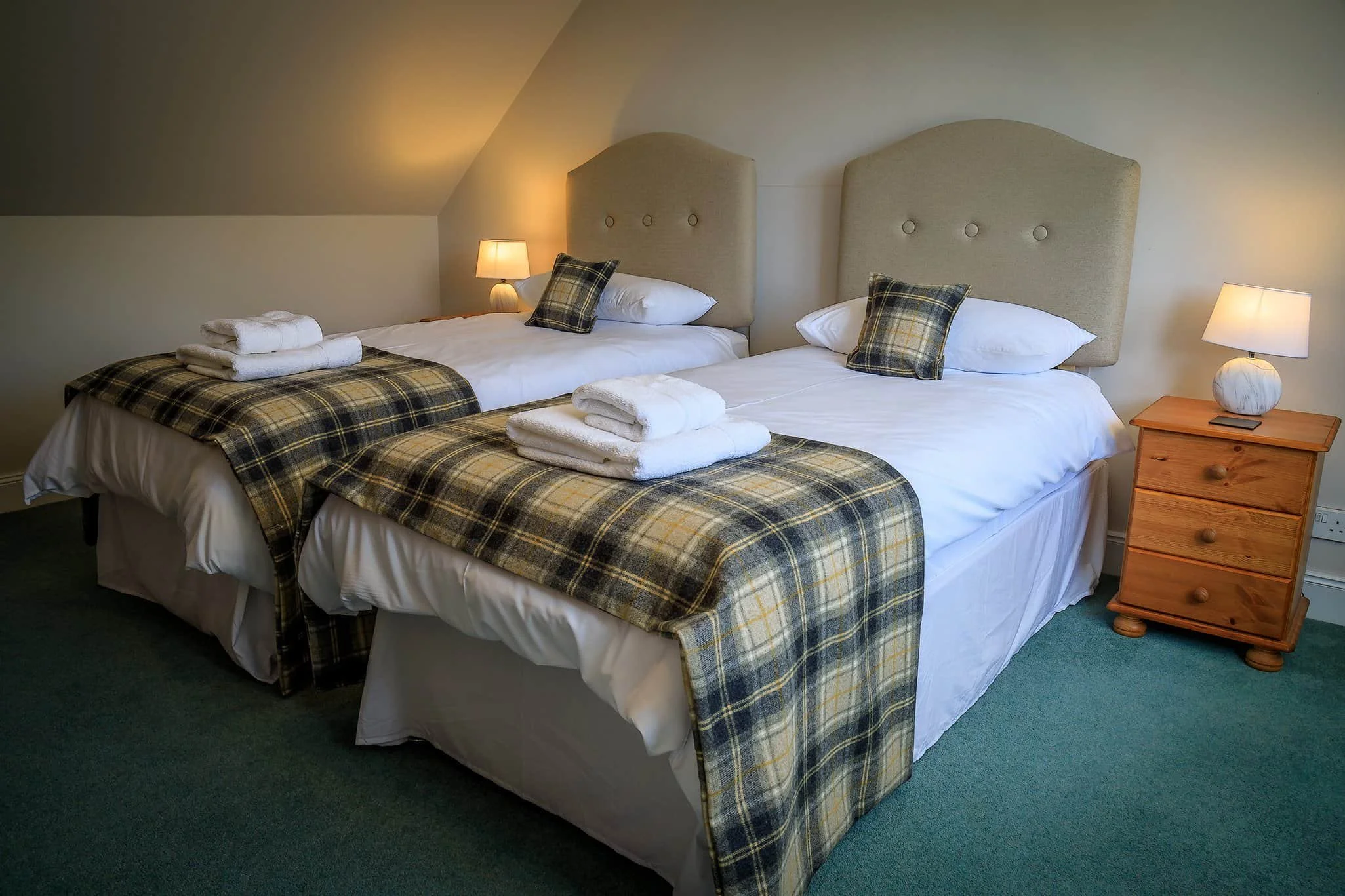 Two twin beds with plaid blankets, pillows, and folded towels, side by side in a cozy room with a nightstand and lamps.