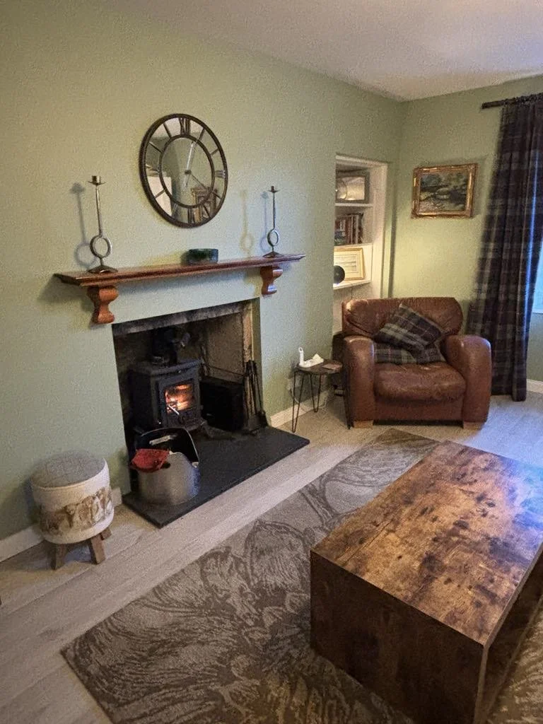 Living room with a fireplace, a leather armchair with a plaid pillow, a wooden coffee table, and a patterned area rug. The wall has a large clock, two candle holders, and a framed painting. There is also a small round ottoman and a side table with a 