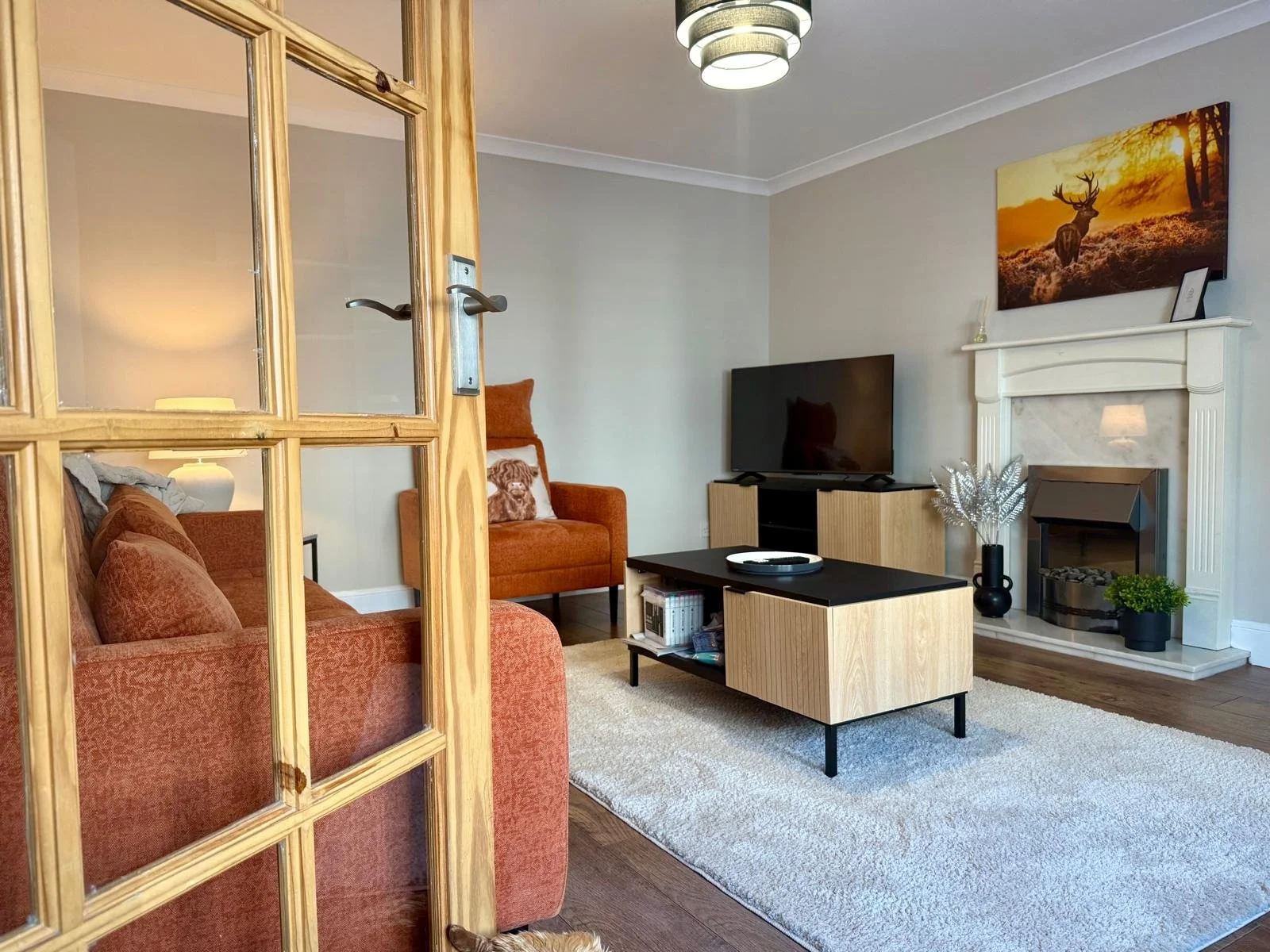 A cozy living room seen through a partially open wooden door with glass panels. The room features two burnt orange sofas, a black and light wood coffee table, a flat-screen TV on a wooden stand, a fireplace with a white mantel, and decorative plants 