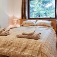 A neatly made bed with beige and white checkered bedding, two rolled towels on top, and a window with brown curtains showing greenery outside. A small lamp is on the nightstand beside the bed.