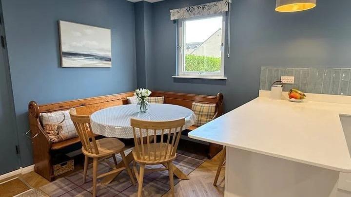 A cozy kitchen nook with a round dining table covered with a white tablecloth, surrounded by wooden chairs and a corner bench with checkered cushions, a vase with flowers on the table, and a window with a valance overlooking a green yard.