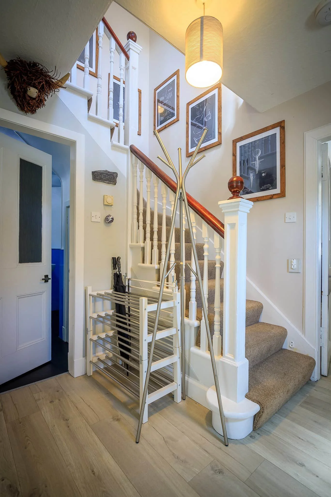 Interior view of a home staircase with framed artwork on the wall, a pendant light hanging from the ceiling, and coat racks and shoes near the entrance.