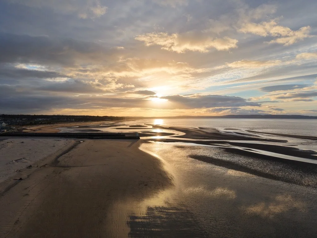Golden sunrise over a sandy beach with tidal pools and dramatic clouds reflecting on the wet sand.