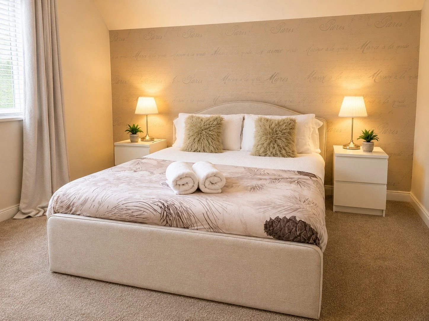 A cozy bedroom with a neatly made bed, two fluffy pillows, and two rolled-up towels on top. There are two nightstands with lamps and small potted plants. Light curtains cover a window on the left.