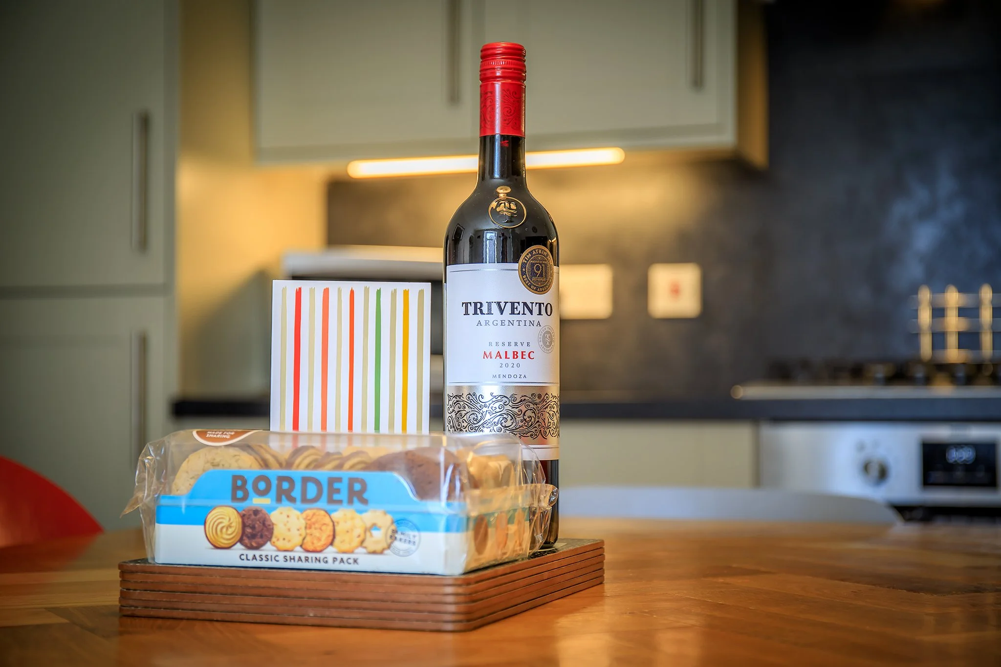 A bottle of Trivento Argentina Reserve Malbec wine, a package of Border cookies, and a colorful striped gift bag on a wooden table in a modern kitchen.