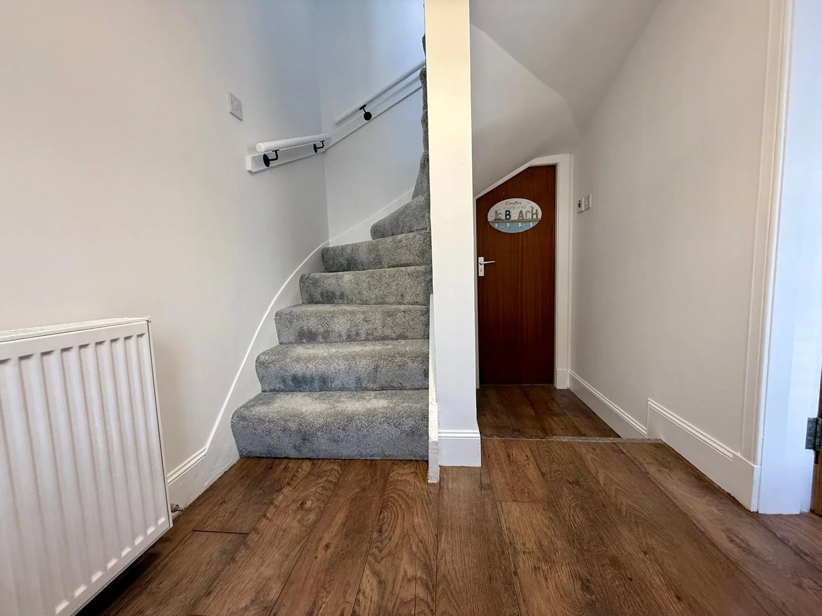 Interior of a house entryway with wooden flooring, a staircase with beige carpet, white walls, and a small wooden door with a beach-themed sign that reads 'Beach' on it.
