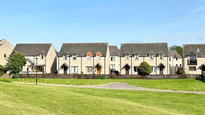 Row of modern beige houses with pitched roofs, small front yards, and a grassy park in the foreground under a clear blue sky.