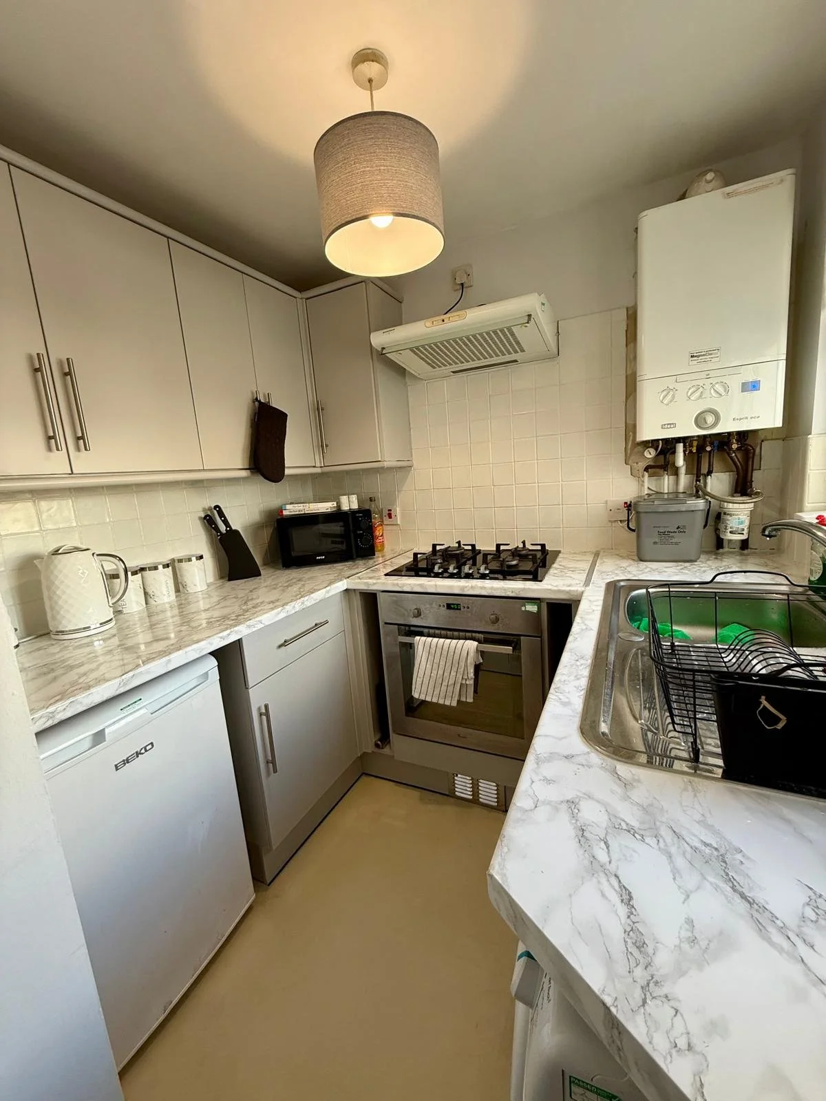 A small kitchen with white cabinets, a marble countertop, a gas stove, a small refrigerator, a microwave, and a sink with a dish rack. There is a ceiling light and a boiler on the wall.