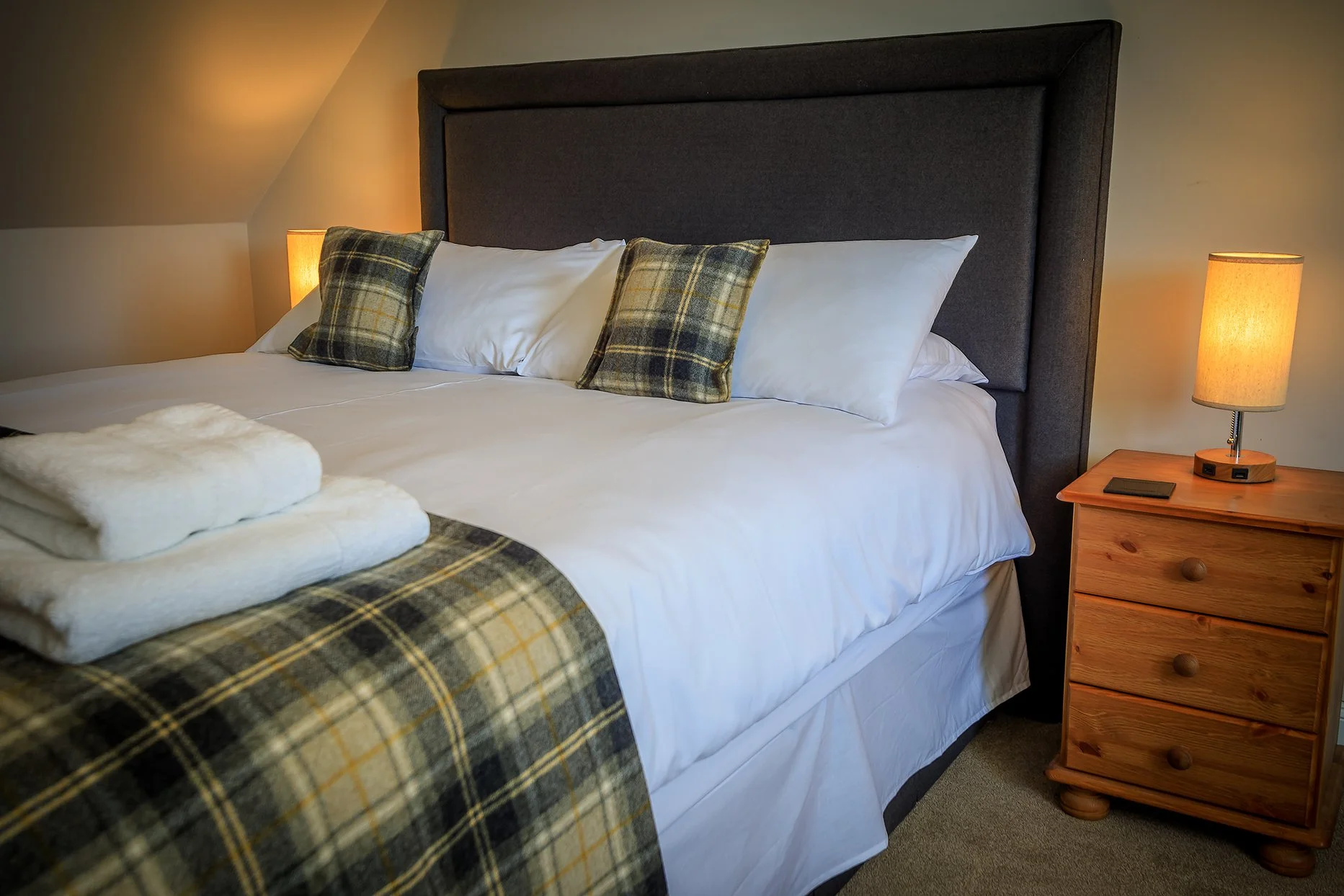 A neatly made bed with white sheets, plaid pillows, and a plaid blanket at the foot. Two folded towels rest on the blanket. A wooden nightstand with a lamp and a remote control are beside the bed.