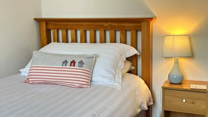 A neatly made bed with white linens, a wooden headboard, and decorative pillows featuring striped and lighthouse designs. A bedside table with a lamp and a remote is beside the bed.