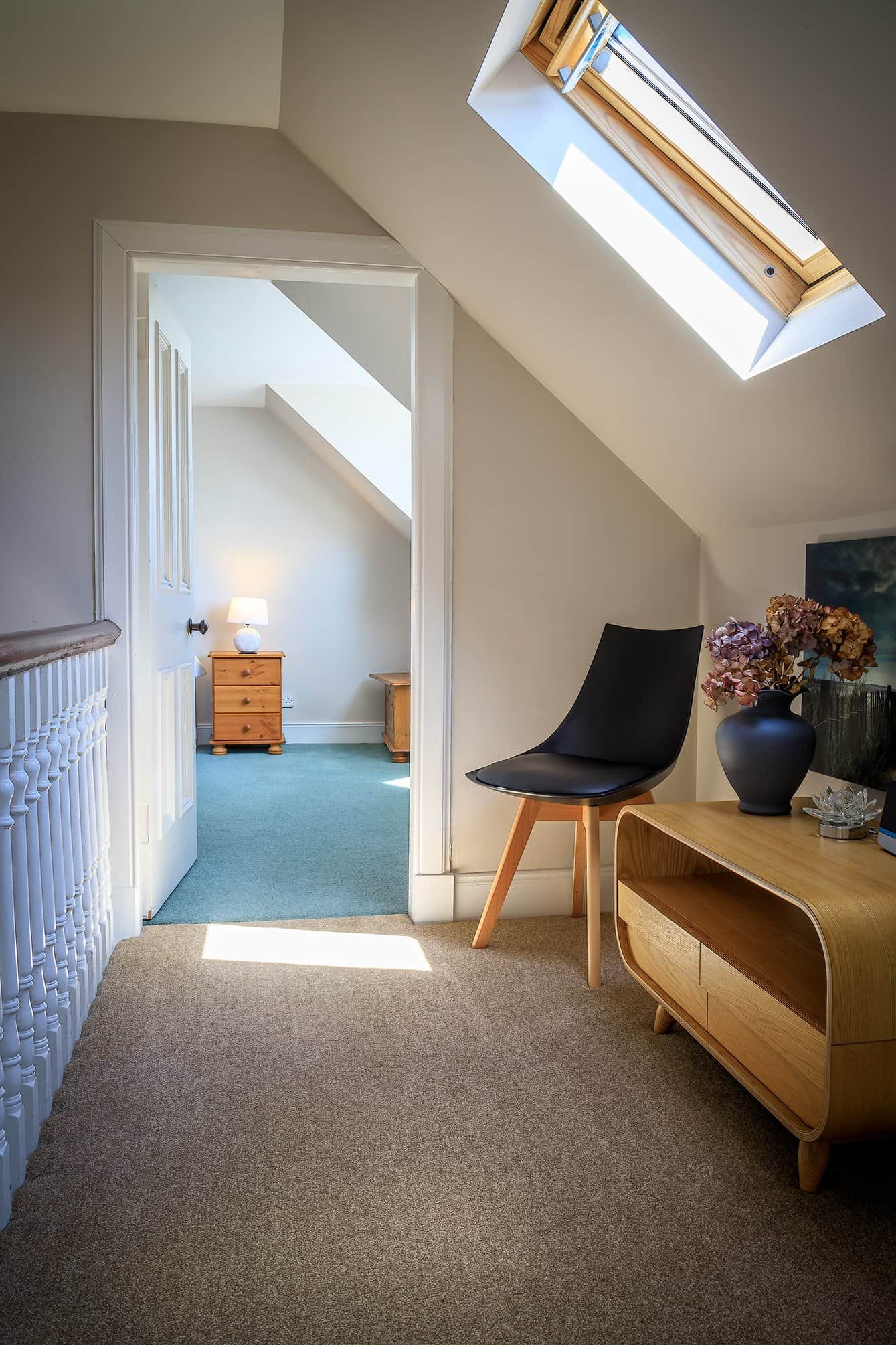 Interior view of a cozy attic space with a skylight, a black chair, a wooden TV stand with a vase of flowers, and a glimpse into a bedroom with a lamp and nightstand.