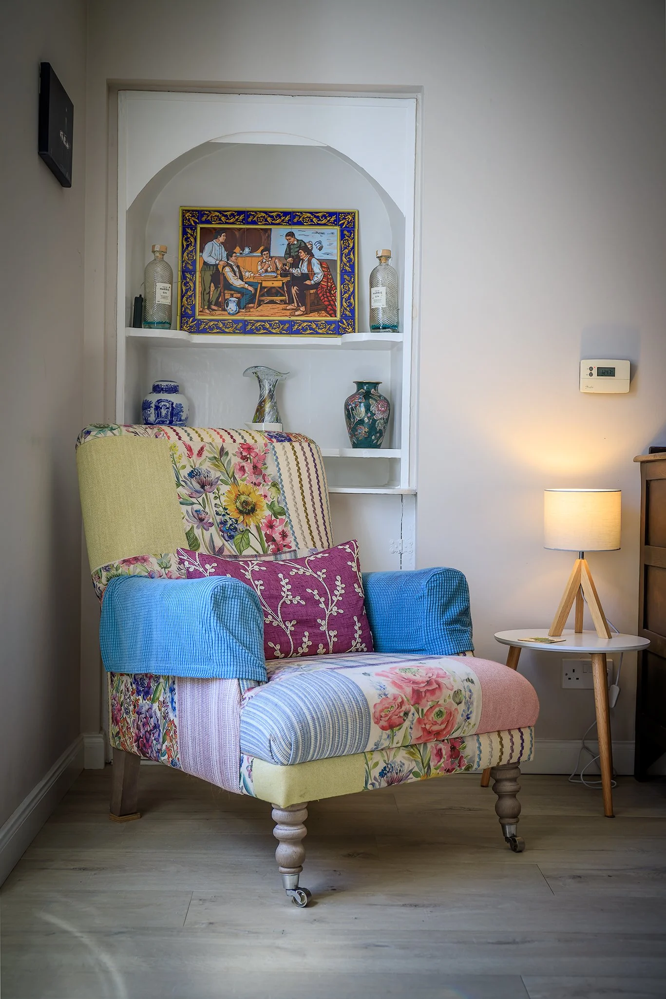 A colorful armchair with floral, striped, and patchwork fabric patterns on a wooden floor next to a white side table with a lamp.