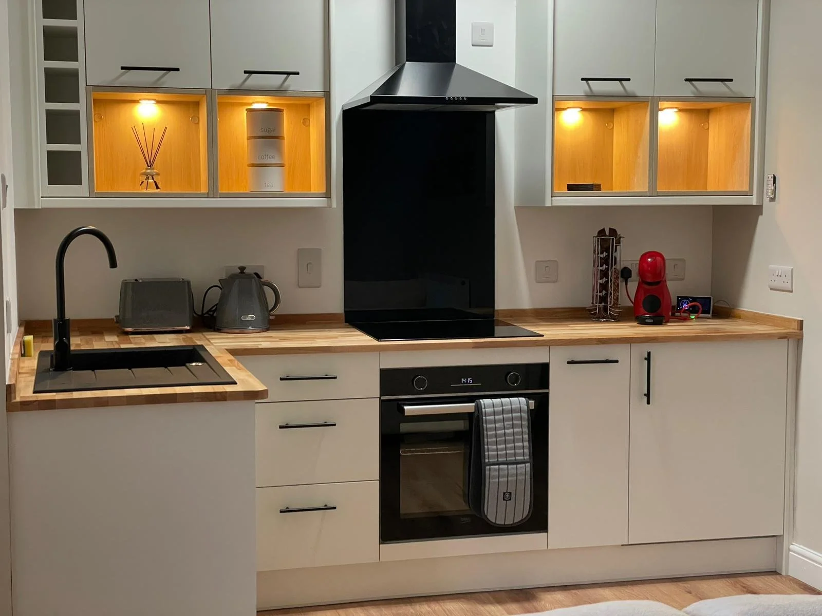 Modern kitchen with white cabinets, wooden countertop, black sink, black oven, and stainless steel appliances. Open shelves and cabinets with warm lighting display decorative jars and kitchen essentials, with a black range hood above the stove.