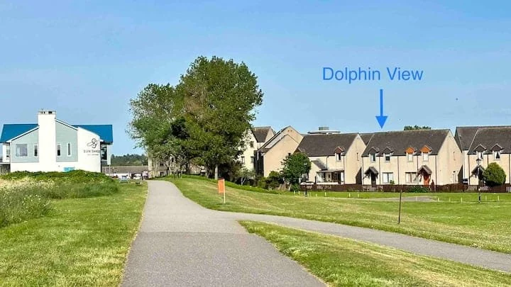 A paved walkway in a grassy park leads towards residential buildings in the distance. A large tree is on the left side, and the sky is clear blue. An arrow points to a building labeled "Dolphin View" in the background.