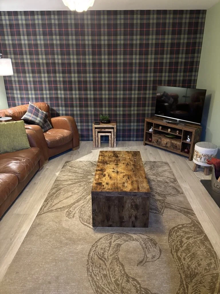 Living room with green walls and plaid wallpaper on the back wall. There is a brown leather sofa with green and plaid pillows, a wooden coffee table, a TV on a wooden stand, and a small side table with a plant. A patterned area rug with floral design