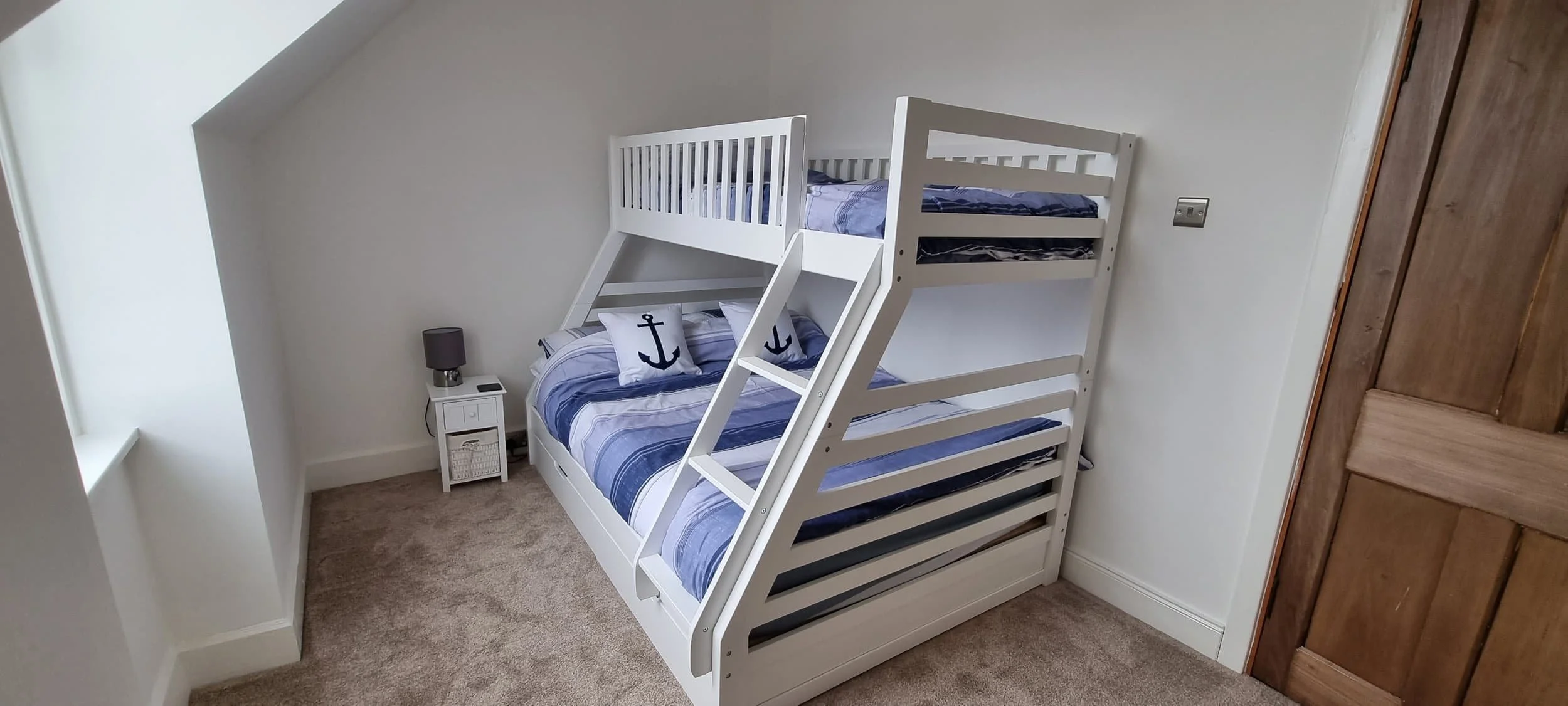 White bed with a bunk bed structure, navy blue and white striped bedding, and pillows with navy anchors, located in a minimalist bedroom with beige carpet, a small white nightstand with a black lamp and a mobile phone, a wooden door, and a window wit