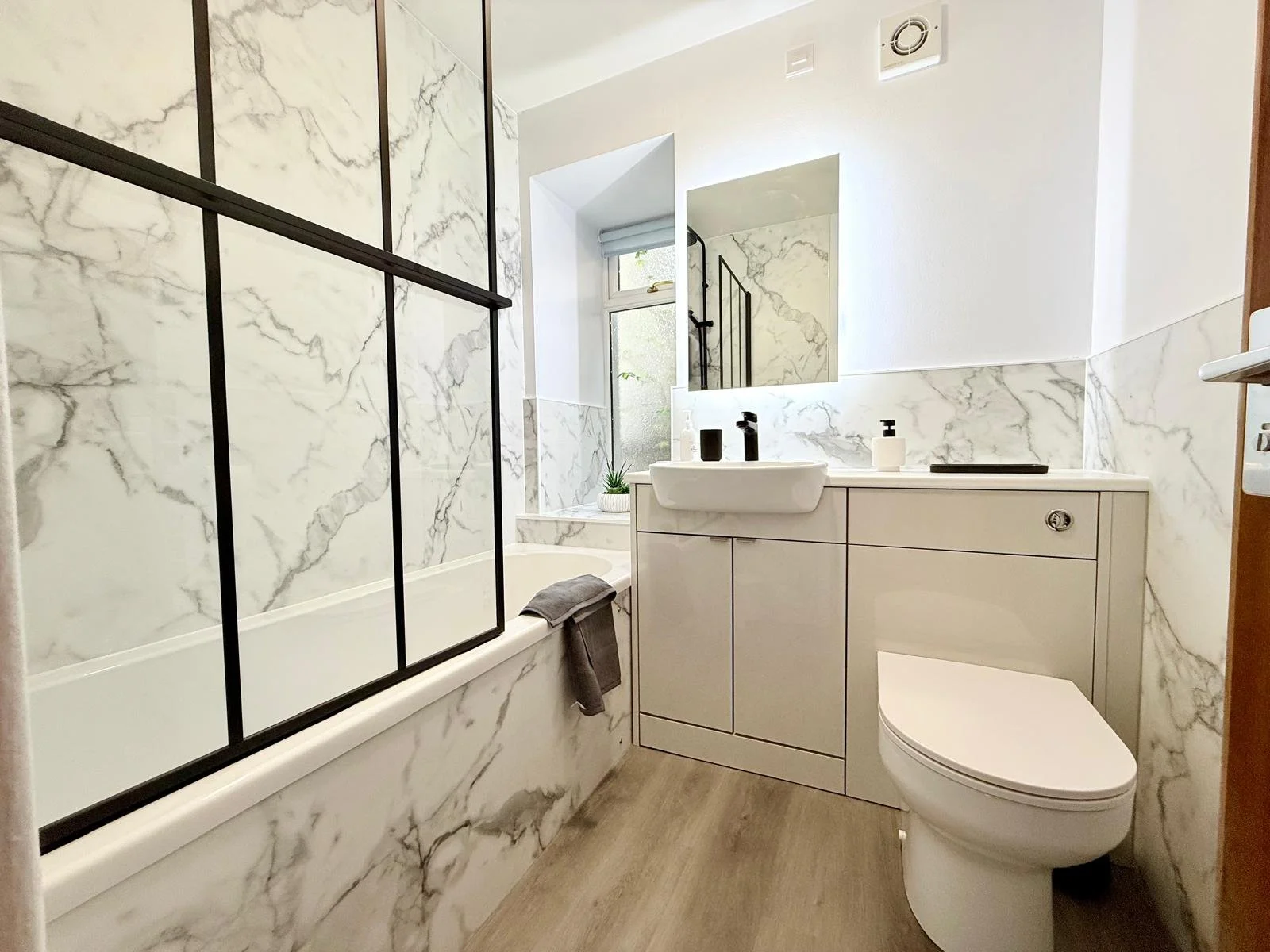 Modern bathroom with marble walls, bathtub with black metal glass partition, white vanity with sink, toilet, small window with blinds, and wooden floor.