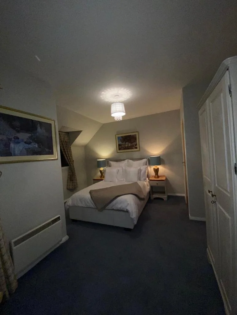 A cozy bedroom with a bed, two matching nightstands with lamps, a framed picture above the bed, and a white wardrobe. The room has soft lighting and curtains on the side window.