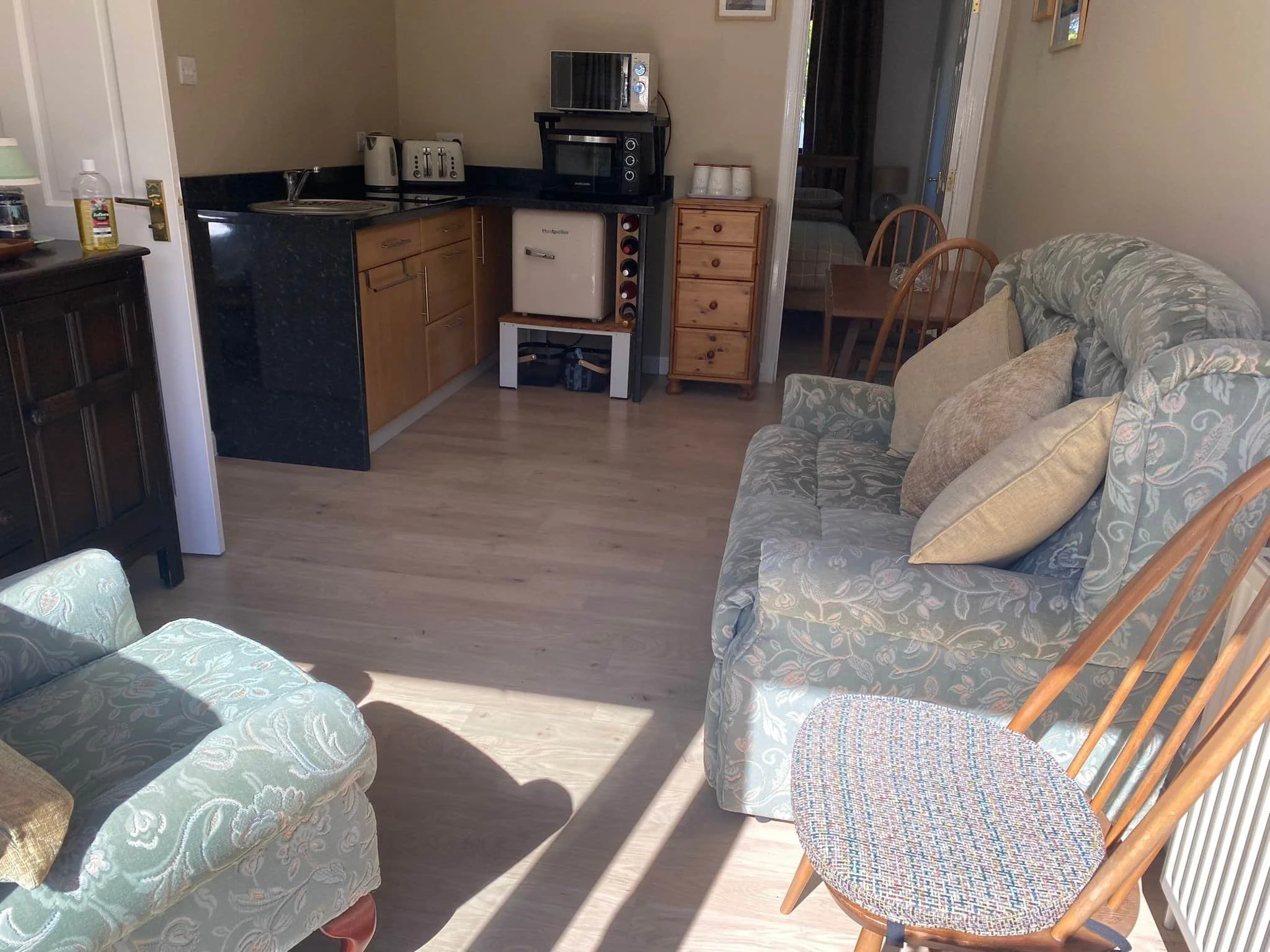 Living room with a sofa and a wooden chair, kitchen area with small appliances, and a glimpse of a bedroom in the background.