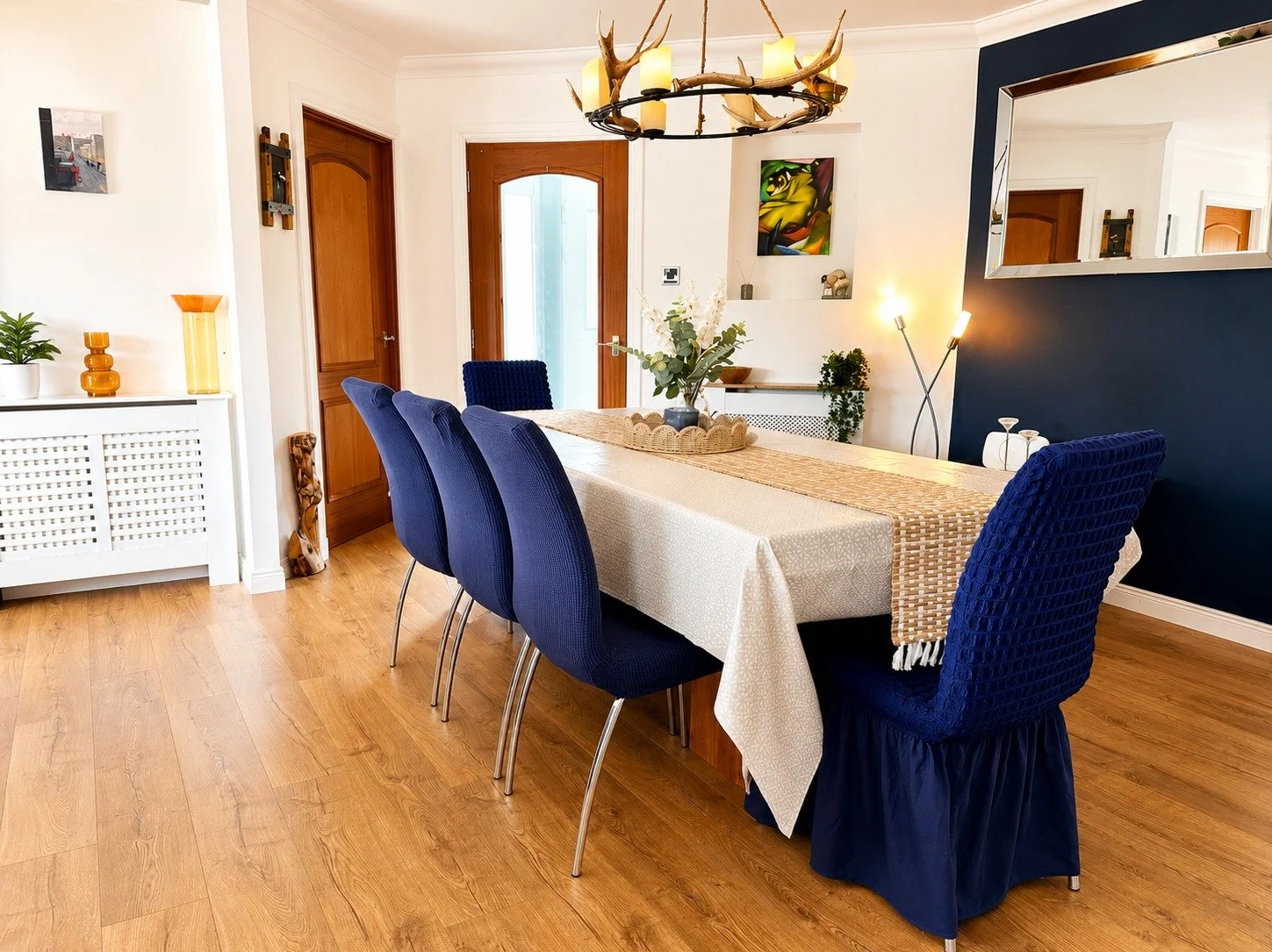 A dining room with a wooden floor, a white radiator cover with decorative items, a wooden door, a mirror on a navy blue wall, a modern floor lamp, and a large table with a beige tablecloth, a tan runner, and a floral centerpiece.