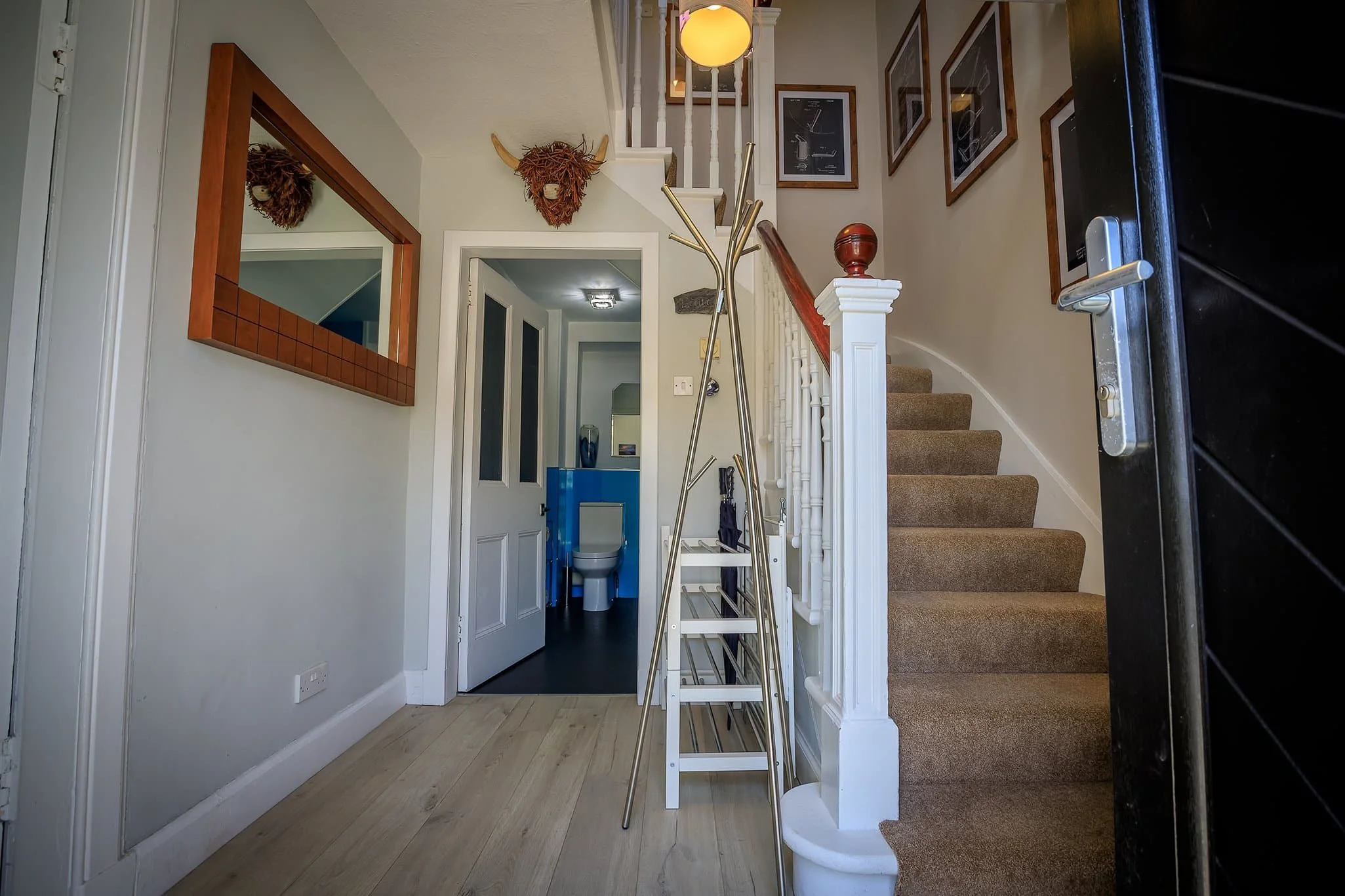 Interior of a house entryway showing stairs with beige carpet, a white railing, a hallway leading to a bathroom, and decorative wall accents including animal head sculptures and framed pictures.