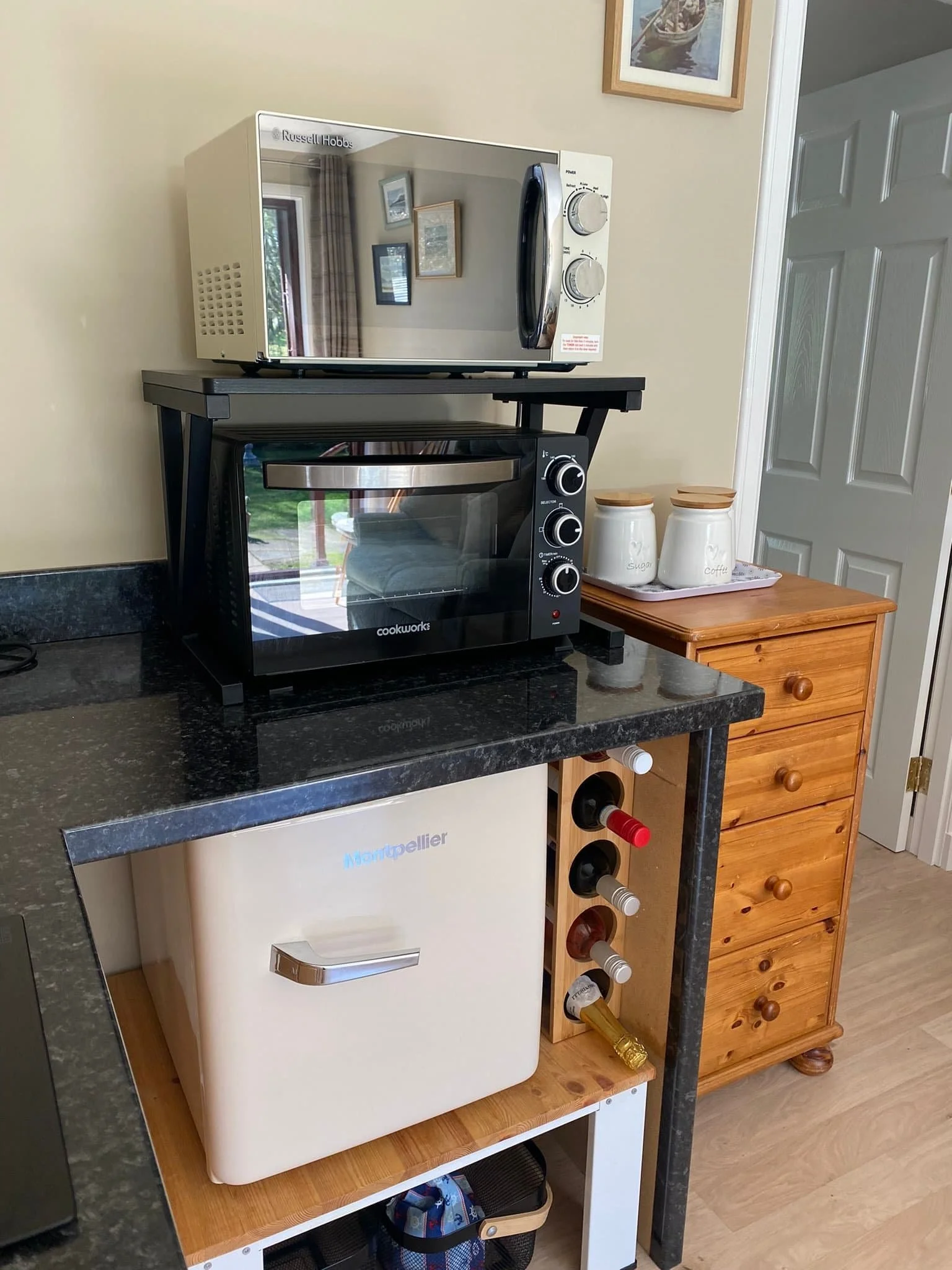 A kitchen counter with a microwave oven on top, a toaster oven below, a small wooden drawer, and a mini fridge underneath. There are ceramic jars labeled 'sugar' and 'coffee' on the drawer. A wine rack with bottles is attached to the side.