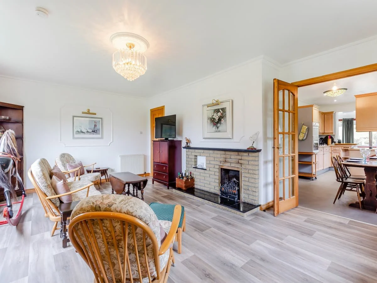 Living room with a beige brick fireplace, wooden furniture, patterned armchairs, a television, and a chandelier, open to a kitchen with wooden cabinets and dining table.